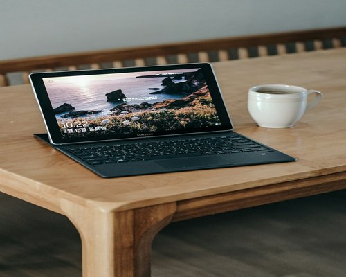 A tablet computer placed neatly on a light gray table surface alongside an open physical notebook and coffee mug during online classes